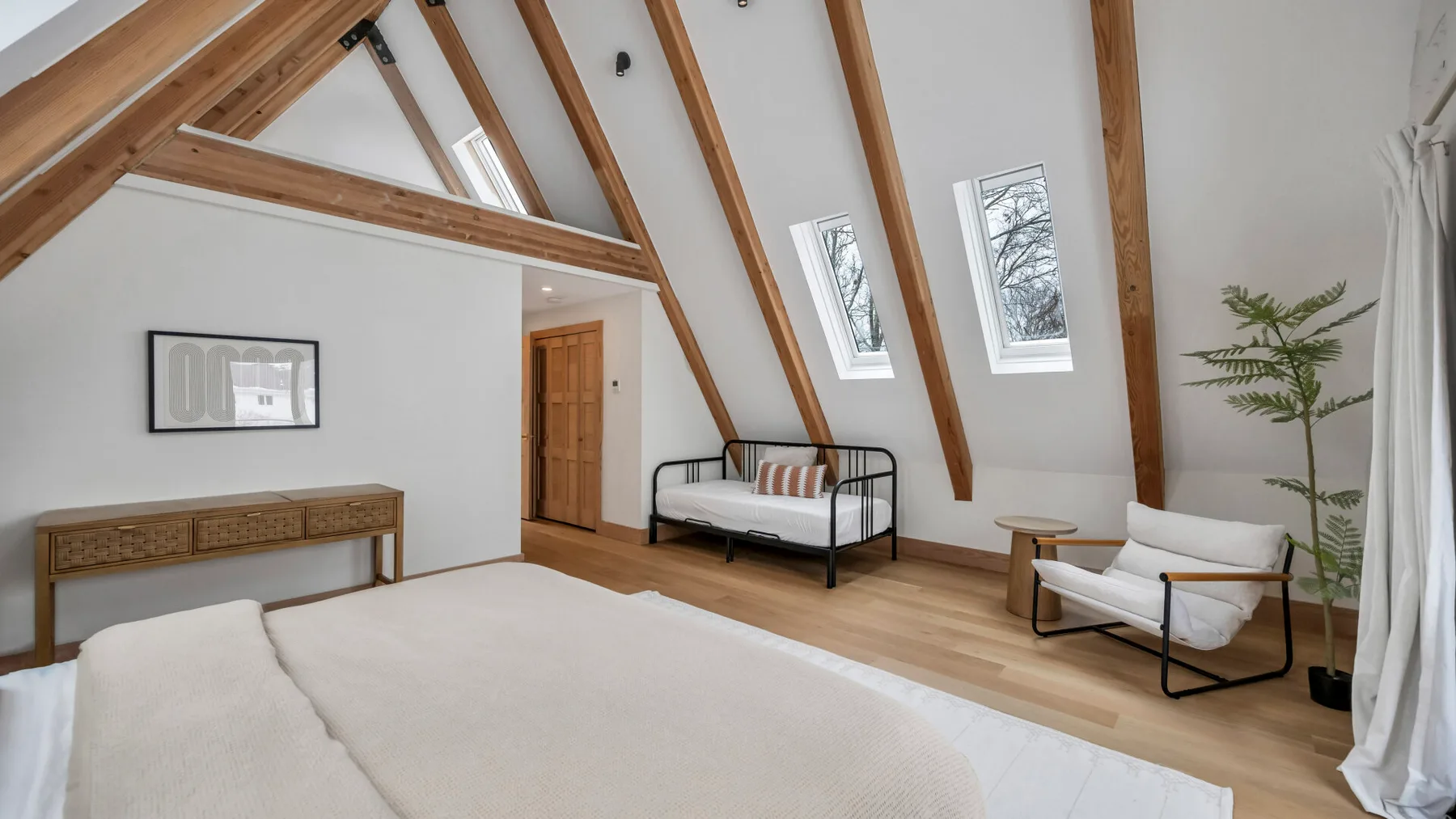 A bright bedroom with a bed, a metal-framed daybed, and sloped timber beams.