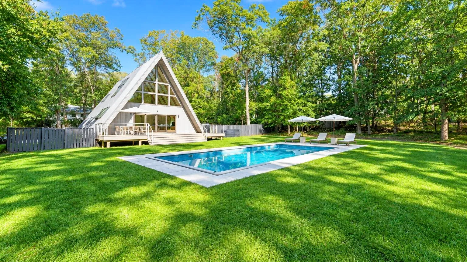 A-frame house and swimming pool set in a grassy, tree-lined yard.