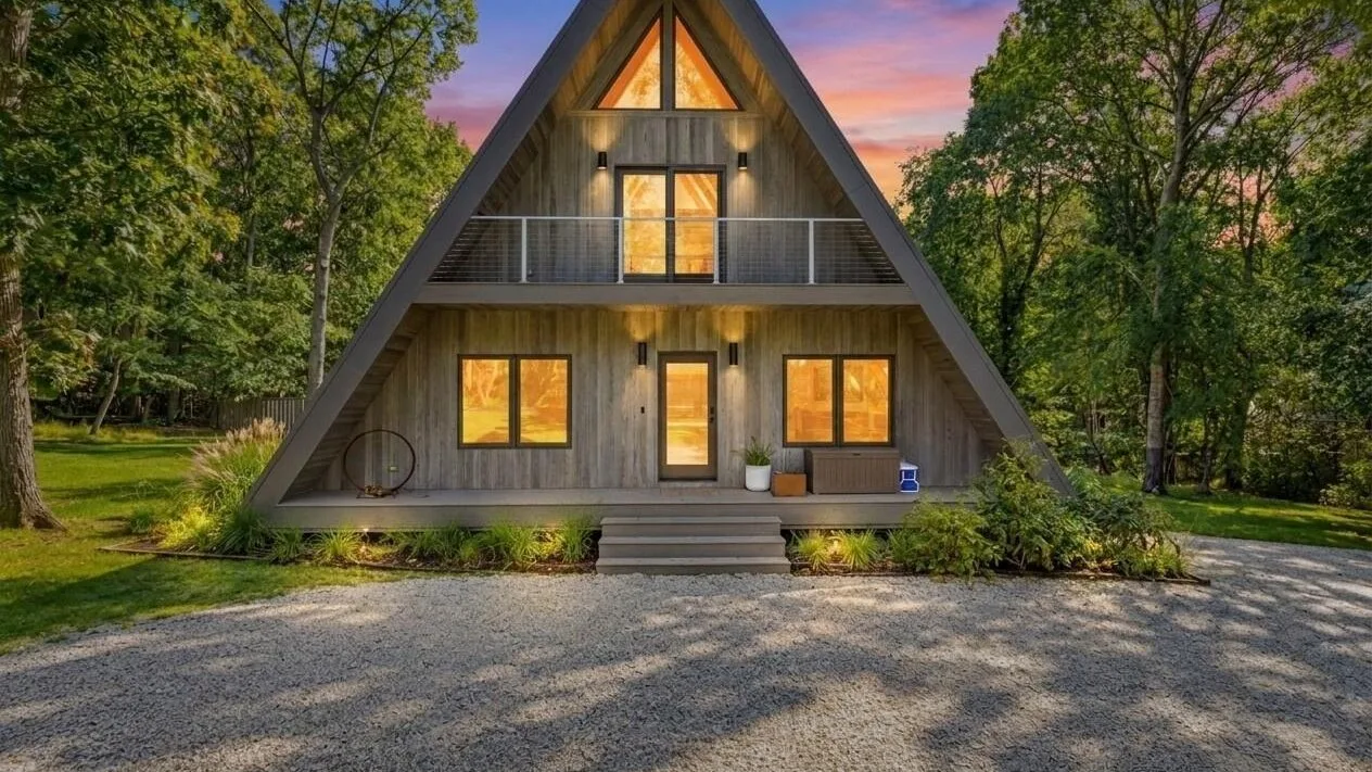 An A-frame house stands among trees beside a gravel driveway at sunset.