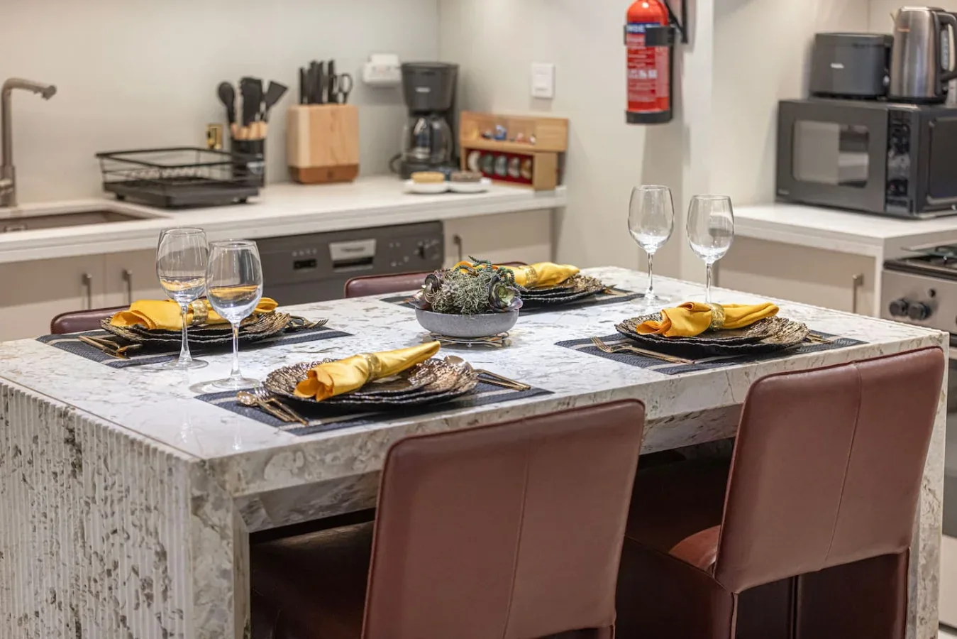 Una mesa de comedor puesta con platos y copas está en una cocina con electrodomésticos sobre la encimera.