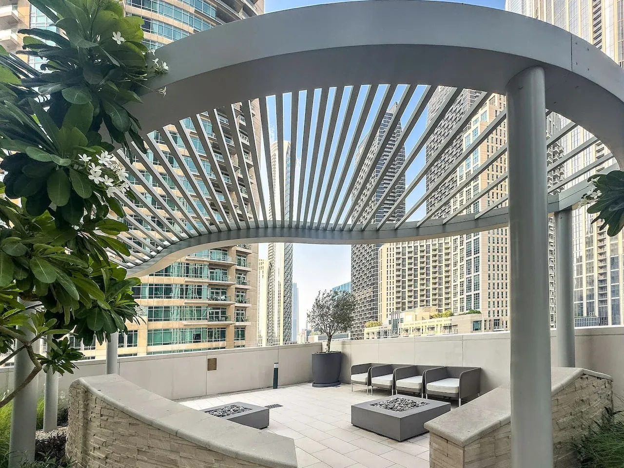 Un patio al aire libre con asientos integrados bajo una pérgola de listones, rodeado de edificios altos.