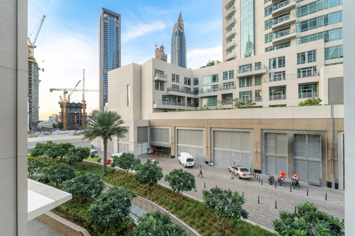 Una calle urbana con edificios modernos altos, grúas de construcción y vegetación ajardinada.