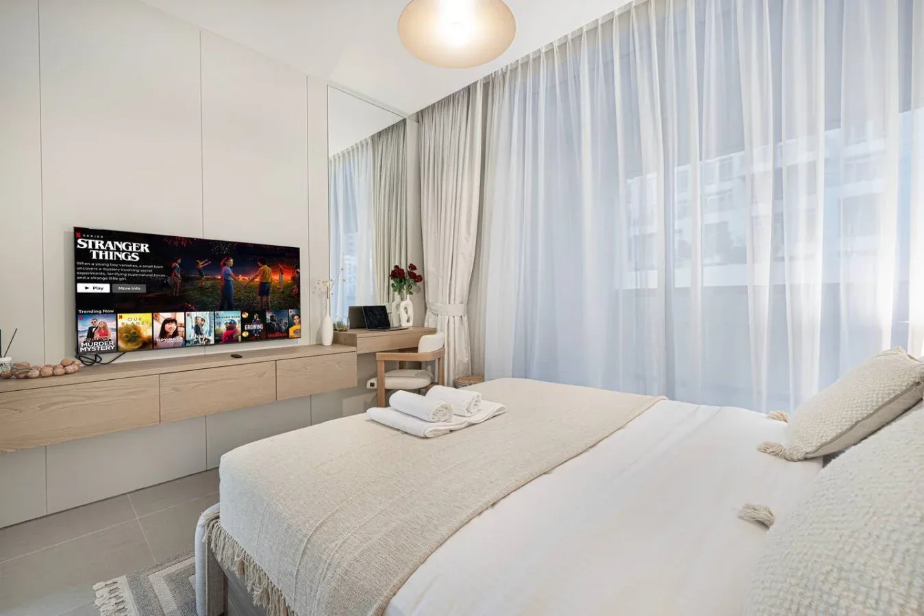 Un dormitorio luminoso con una cama tendida junto a grandes ventanas con cortinas.