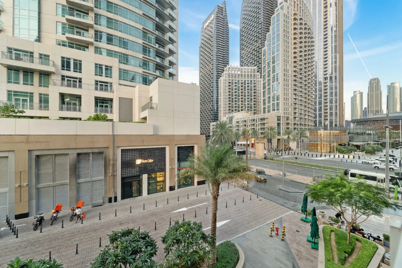 Una plaza peatonal pavimentada con zonas verdes está frente a altos edificios urbanos.