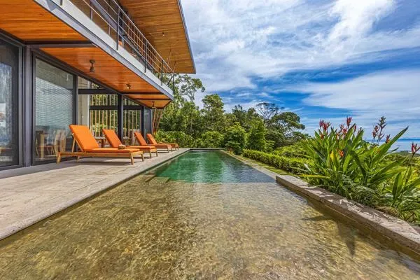 Una villa moderna da a una piscina alargada rodeada de plantas tropicales.