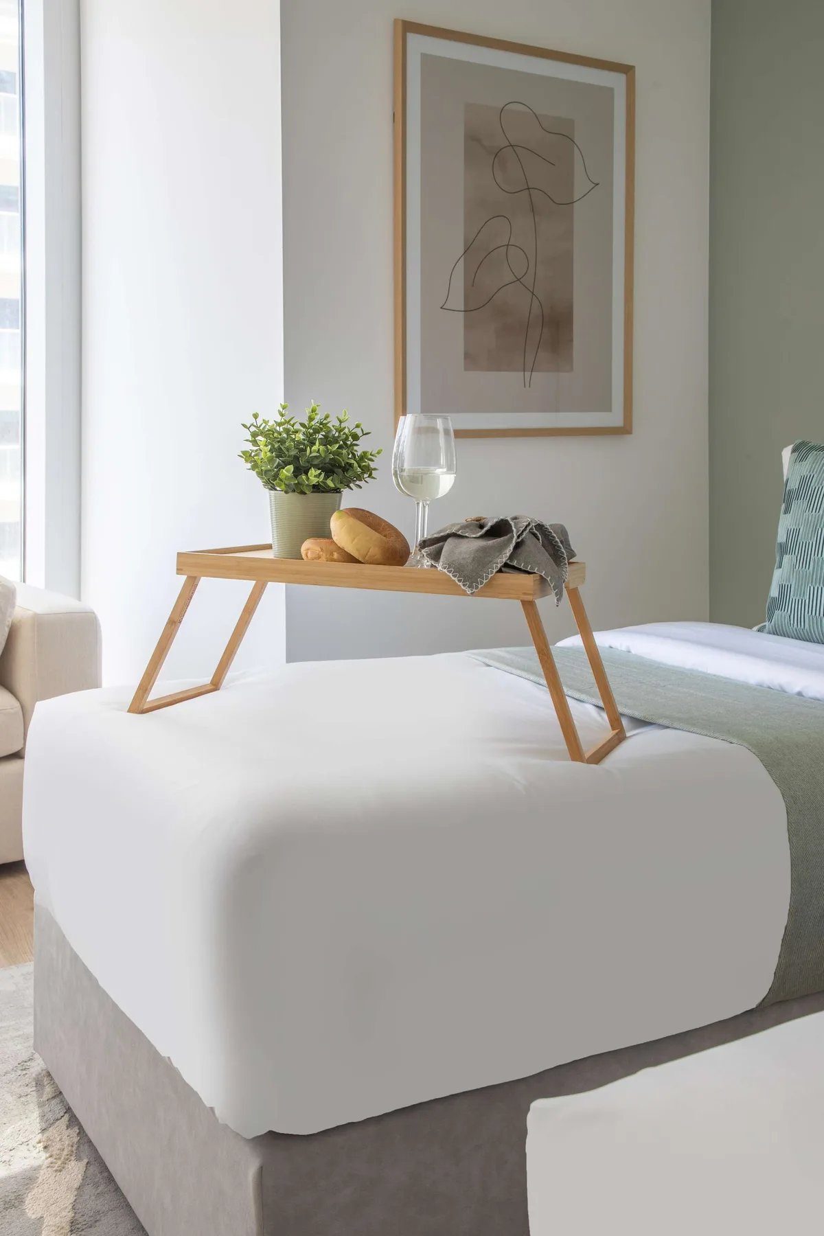 Un dormitorio con una cama blanca y una bandeja de madera con una planta y una copa.