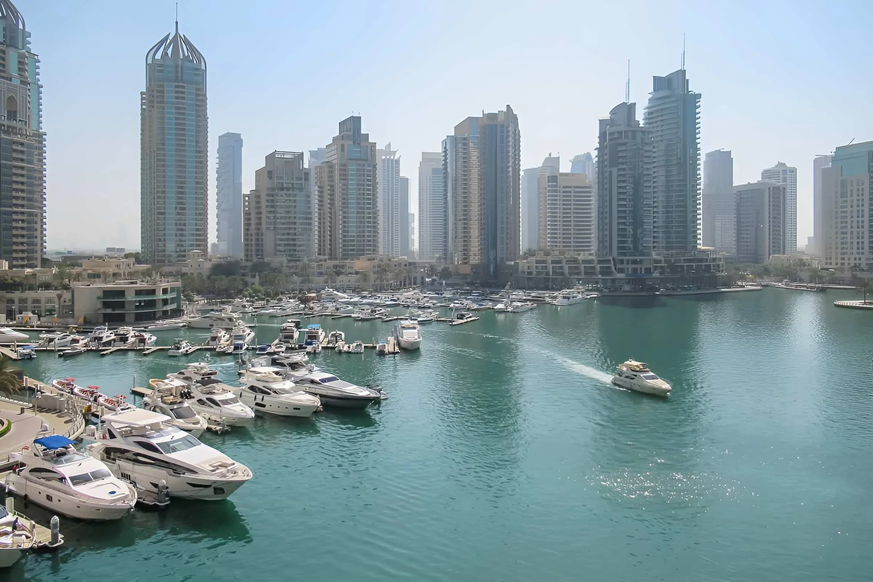 Una marina con barcos sobre aguas turquesas y edificios altos junto al paseo marítimo.