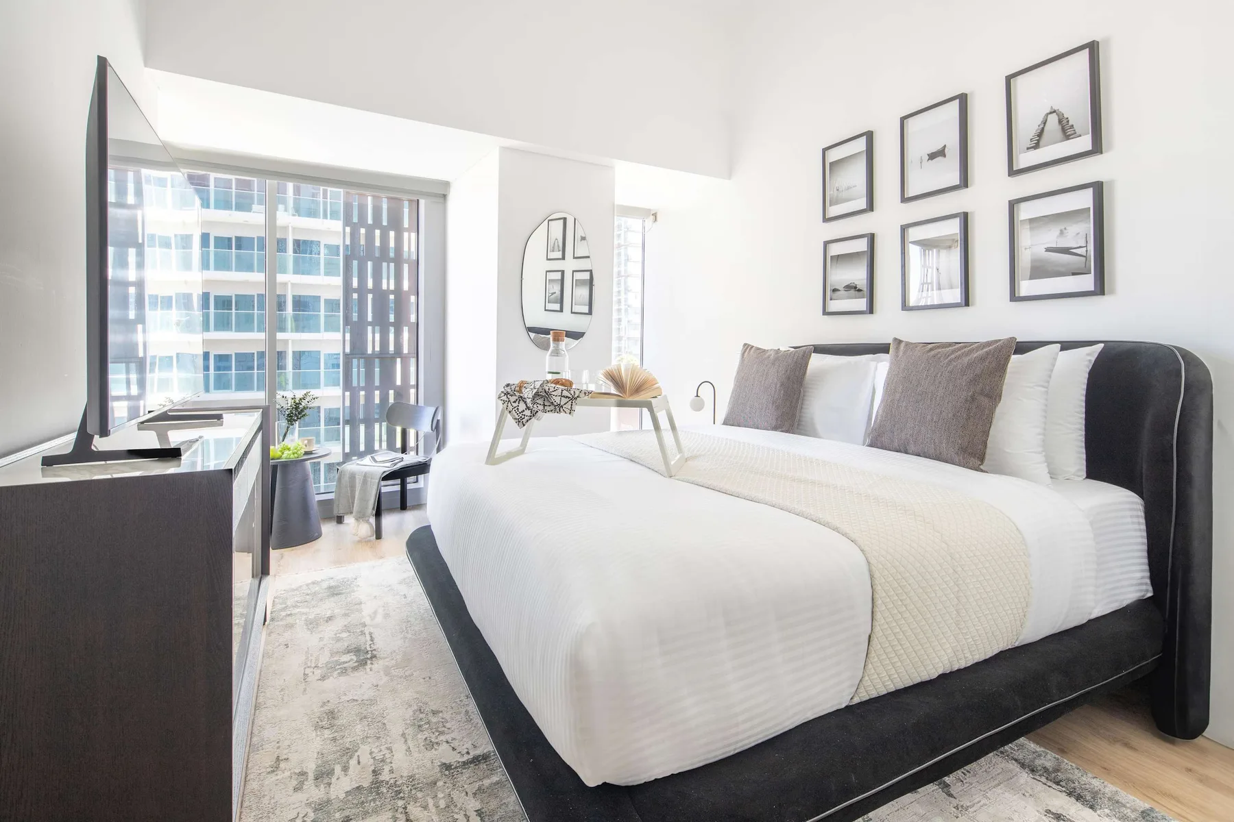 Un dormitorio luminoso con una cama grande, cuadros enmarcados y vistas a la ciudad por la ventana.