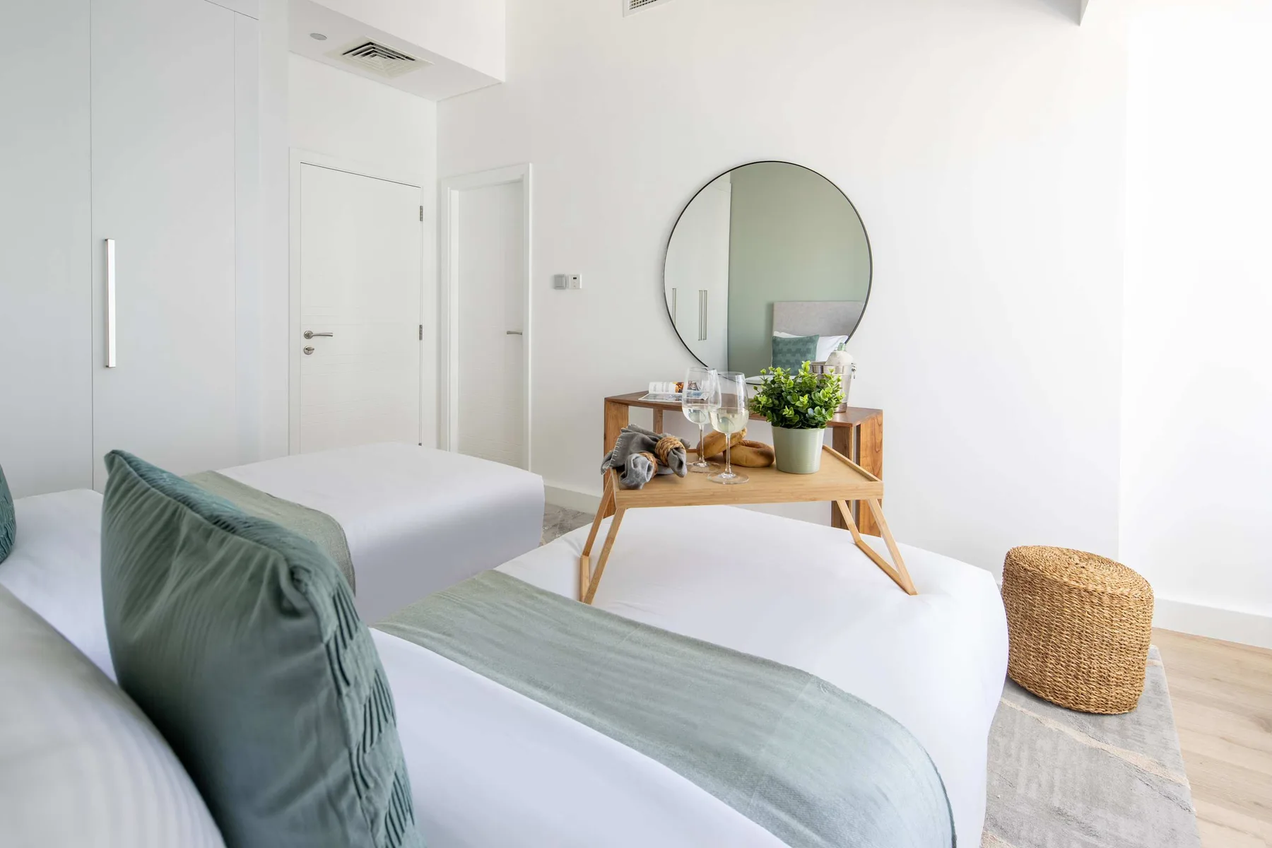 Un dormitorio luminoso con ropa de cama blanca, cojines verdes y un espejo redondo.