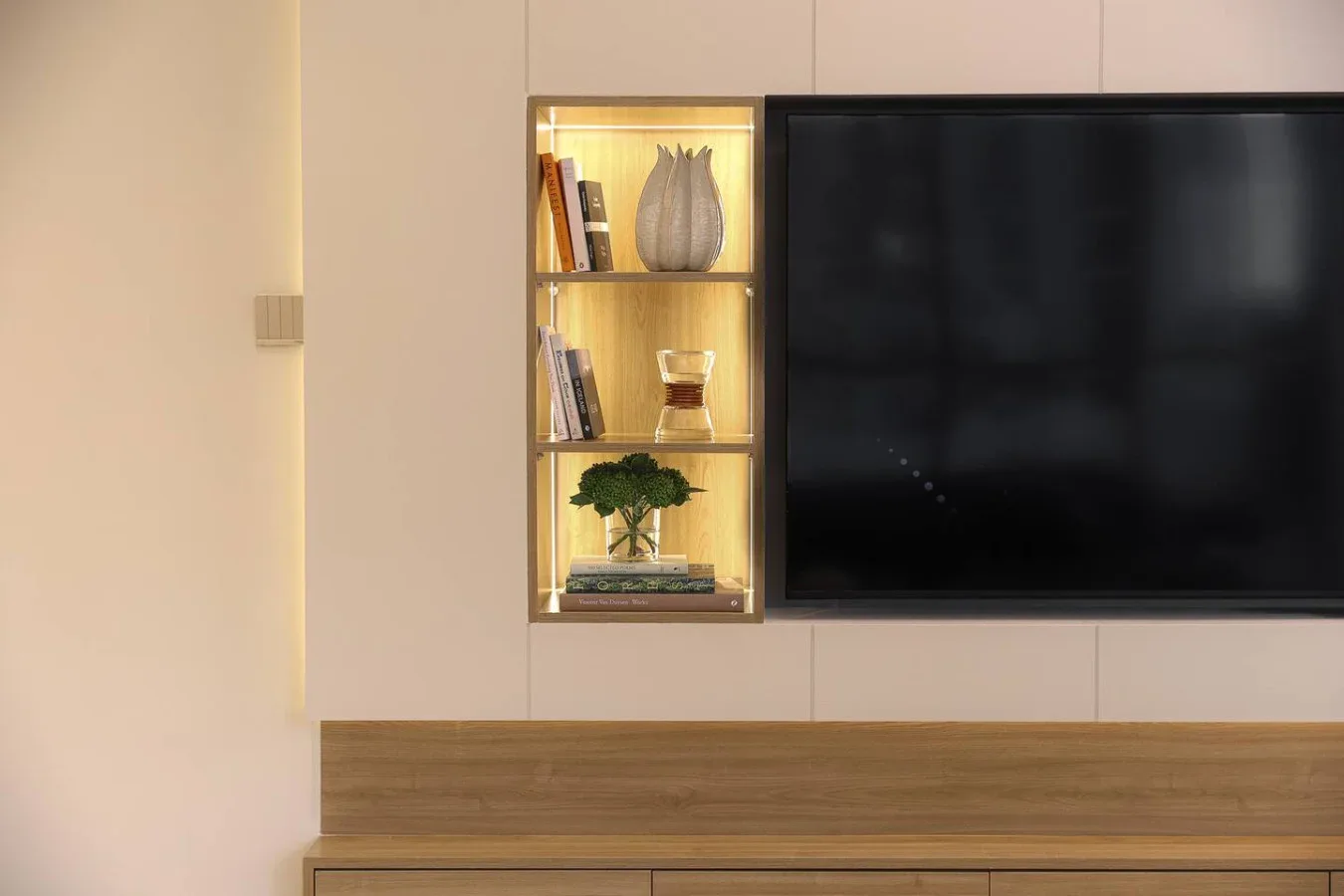 Un televisor montado en la pared está junto a una estantería iluminada, sobre un mueble de madera.