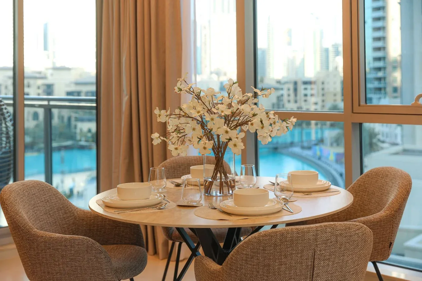 Una mesa de comedor con cubiertos y flores está junto a grandes ventanales.