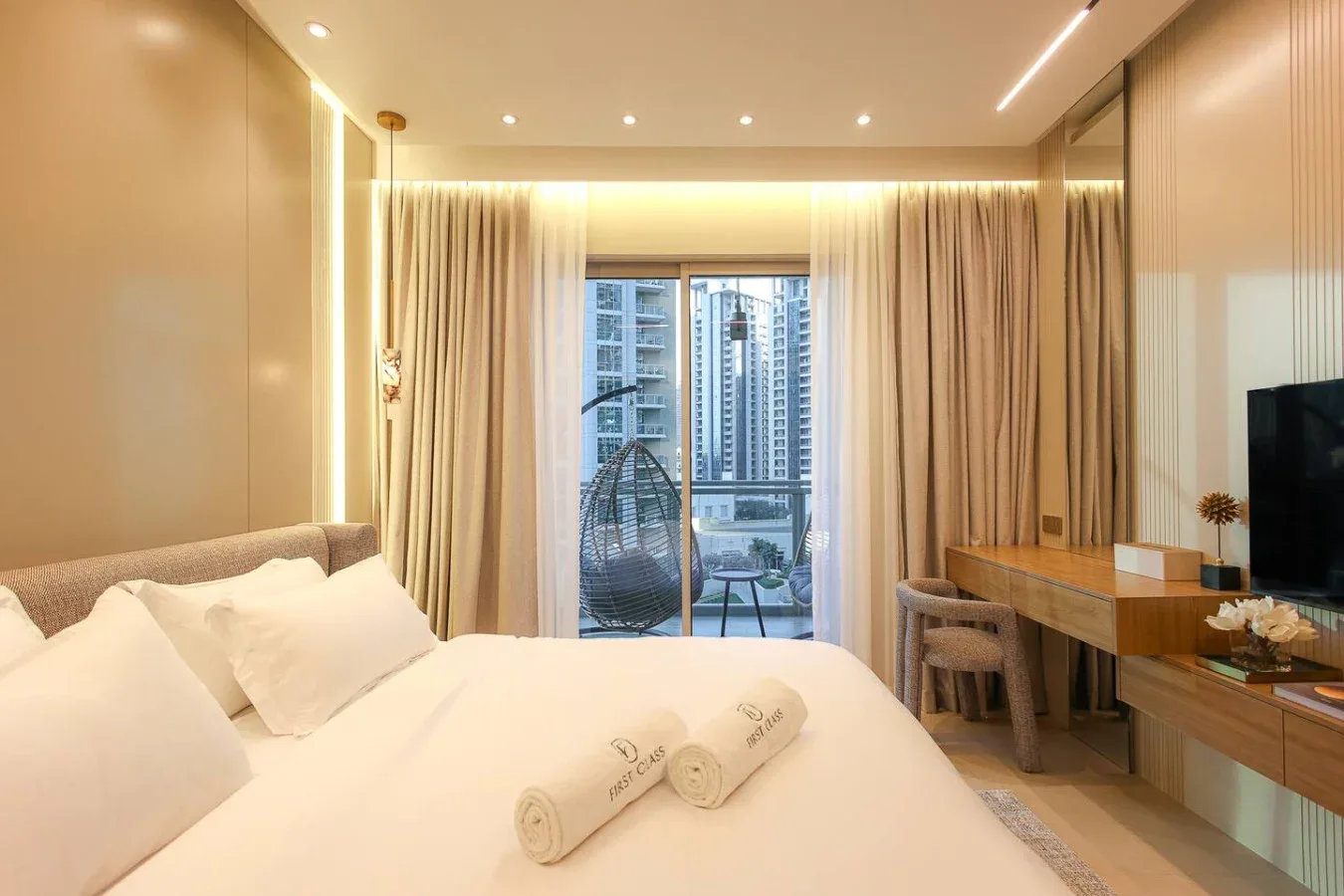 Un dormitorio luminoso con una cama ordenada y una gran ventana con vista a edificios cercanos.