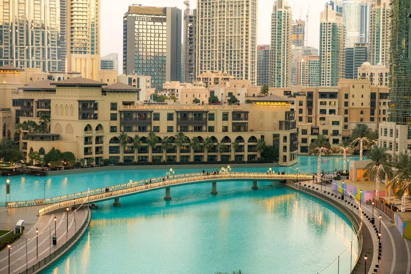 Una gran piscina turquesa está bajo un puente peatonal, con edificios de la ciudad detrás.