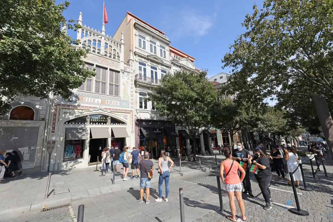 A city street with pedestrians passing storefronts beneath leafy trees.