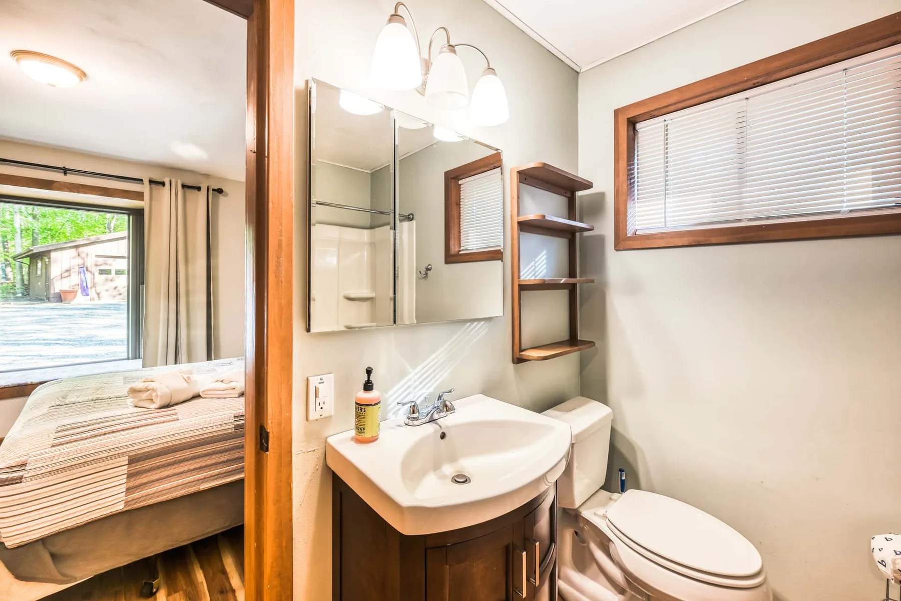 Un baño con lavabo, espejo, inodoro y vista al dormitorio contiguo.