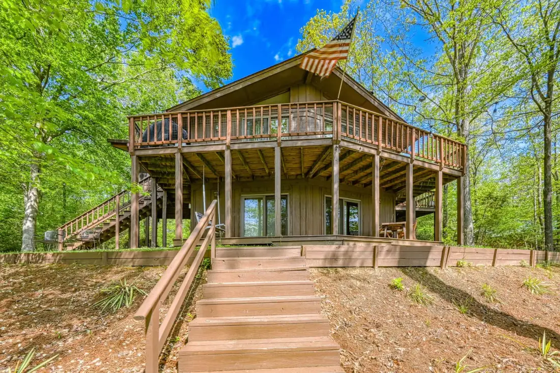 A raised wooden cabin surrounded by trees, with a broad deck and stairs.