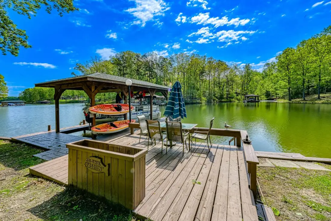 A wooden lakeside deck with outdoor seating and boats beside the water.