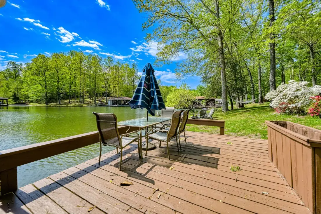 A lakeside deck has outdoor seating and a closed umbrella beside the water.