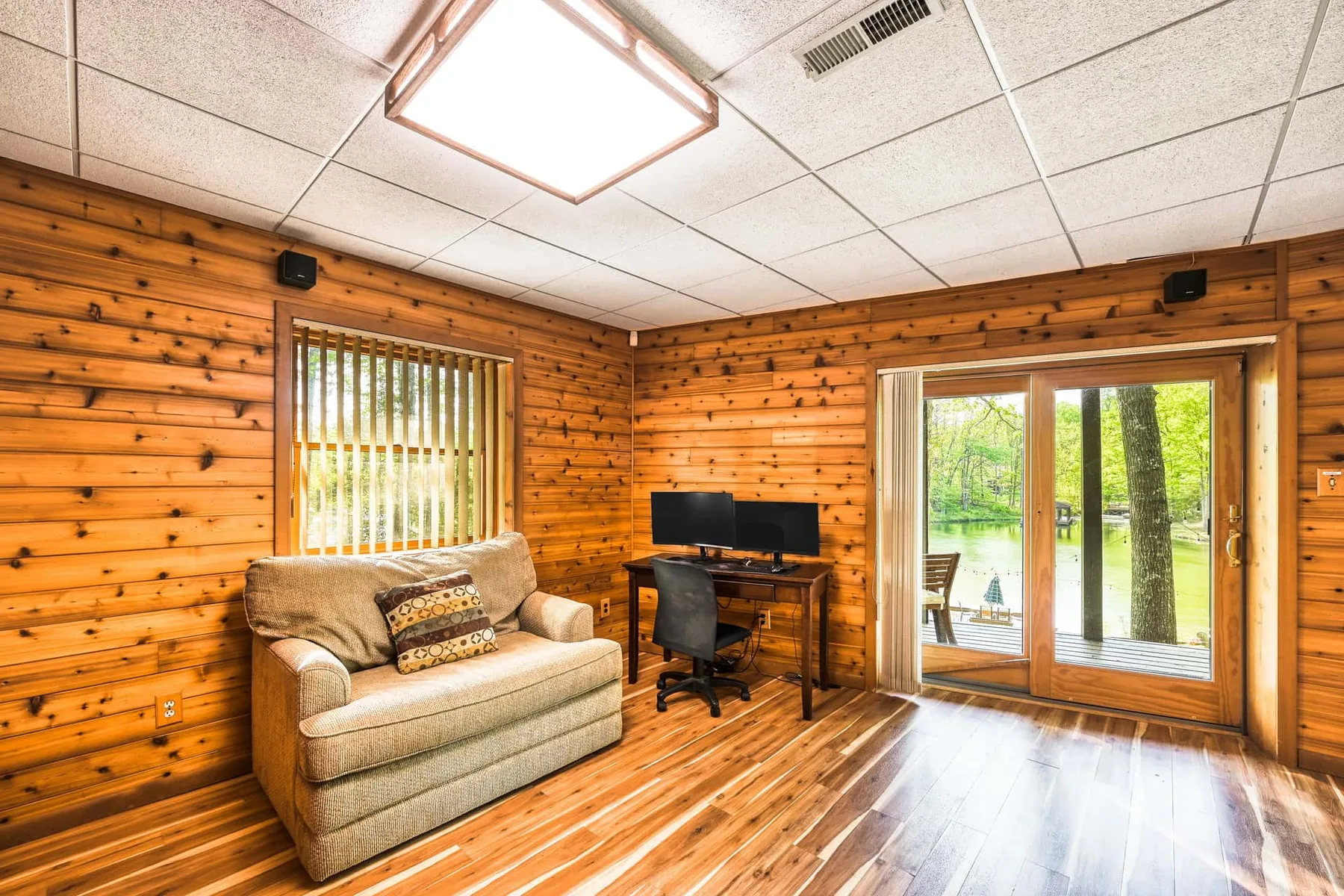 Una habitación revestida de madera tiene un sofá y un escritorio junto a puertas de vidrio que dan a la vegetación.