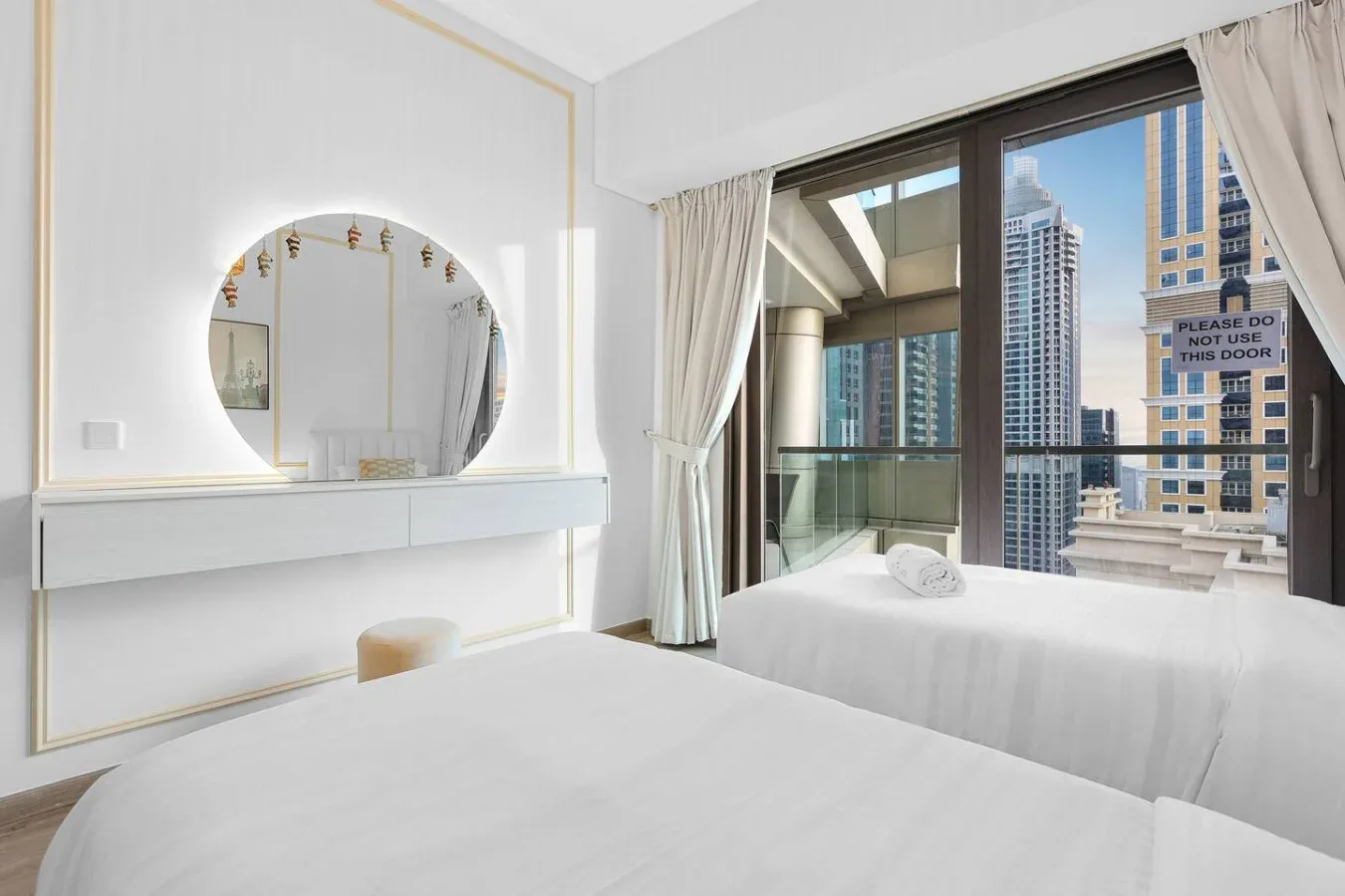 Un dormitorio luminoso con camas junto a un gran ventanal con vistas a edificios de la ciudad.