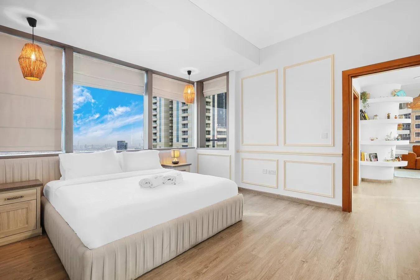 Un dormitorio luminoso con una cama grande junto a amplias ventanas con vistas a edificios cercanos.