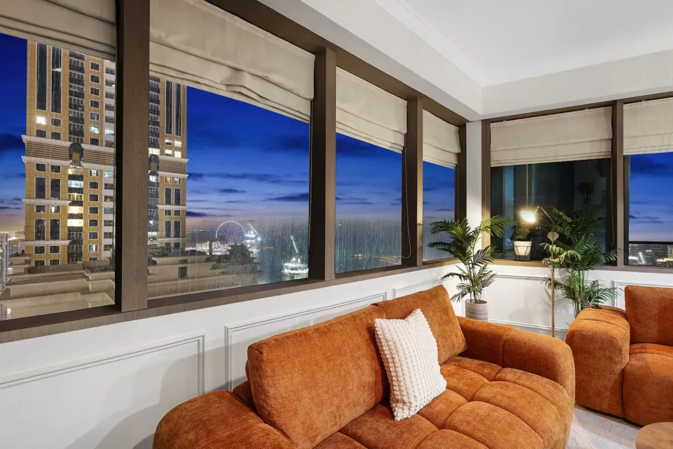 Una sala de estar con asientos naranjas junto a grandes ventanas con vistas a la ciudad al anochecer.