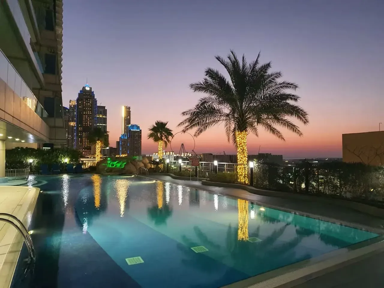 Una piscina en la azotea refleja palmeras y luces de la ciudad al anochecer.