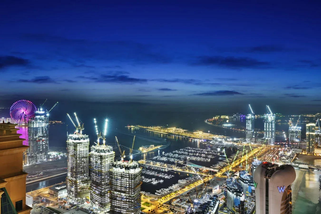 Un horizonte urbano nocturno domina una zona costera con edificios iluminados y grúas.