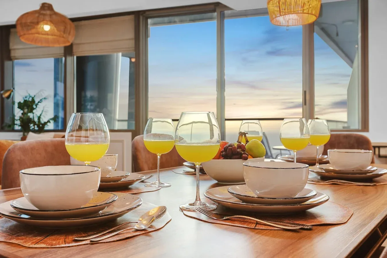 Una mesa de comedor está preparada con platos y copas junto a grandes ventanas.