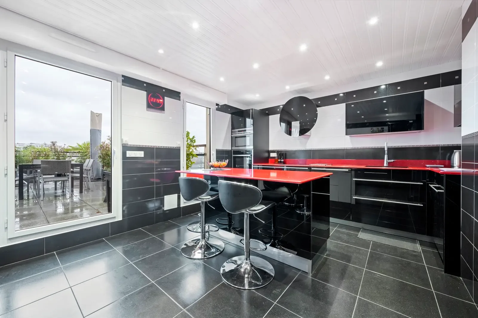 A modern kitchen with a red counter and bar stools beside glass doors.