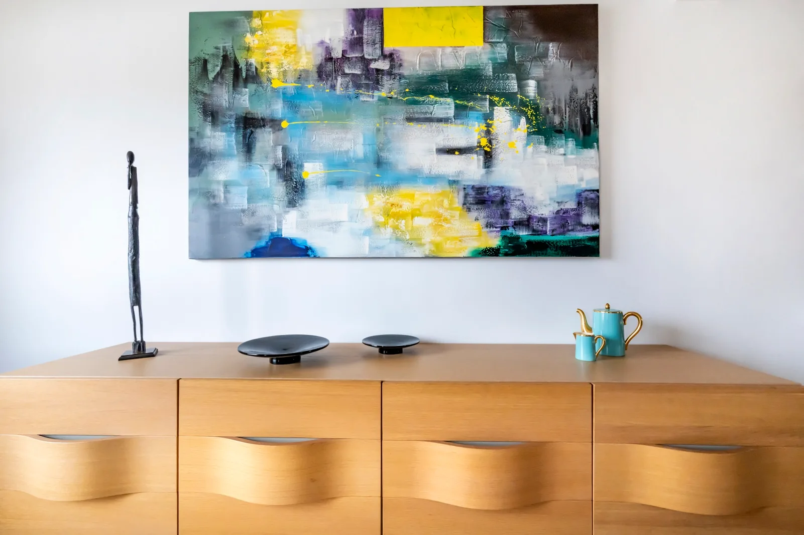 A wooden cabinet displays an abstract painting, a small sculpture, and decorative vessels.