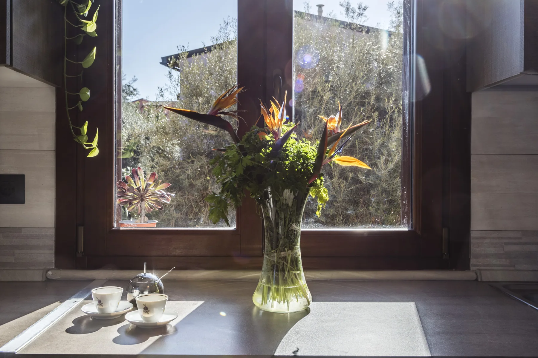 Un alféizar interior iluminado por el sol tiene un jarrón de vidrio con flores naranjas y un juego de té.