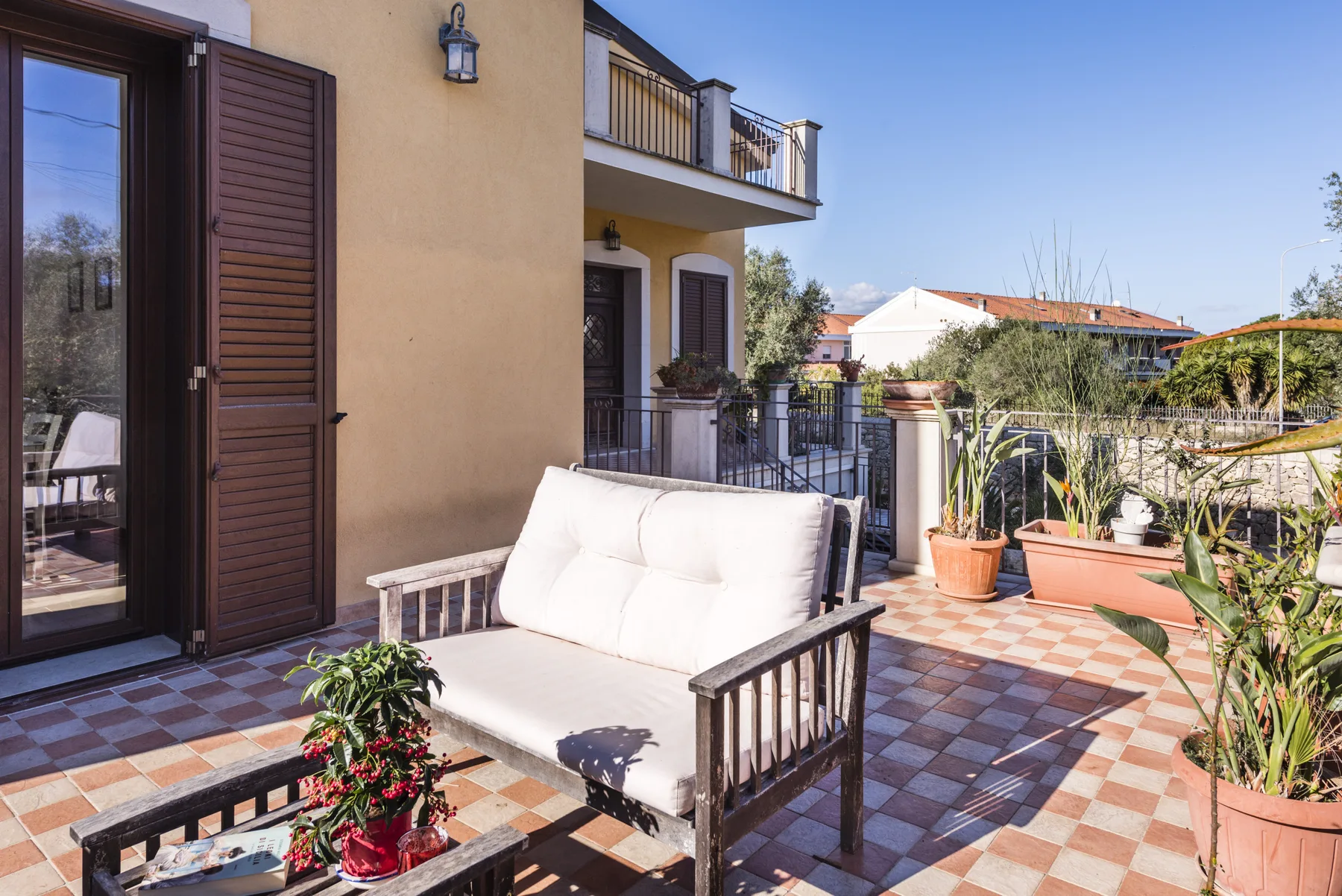 Una terraza exterior con suelo de baldosas, asientos acolchados y plantas en macetas.