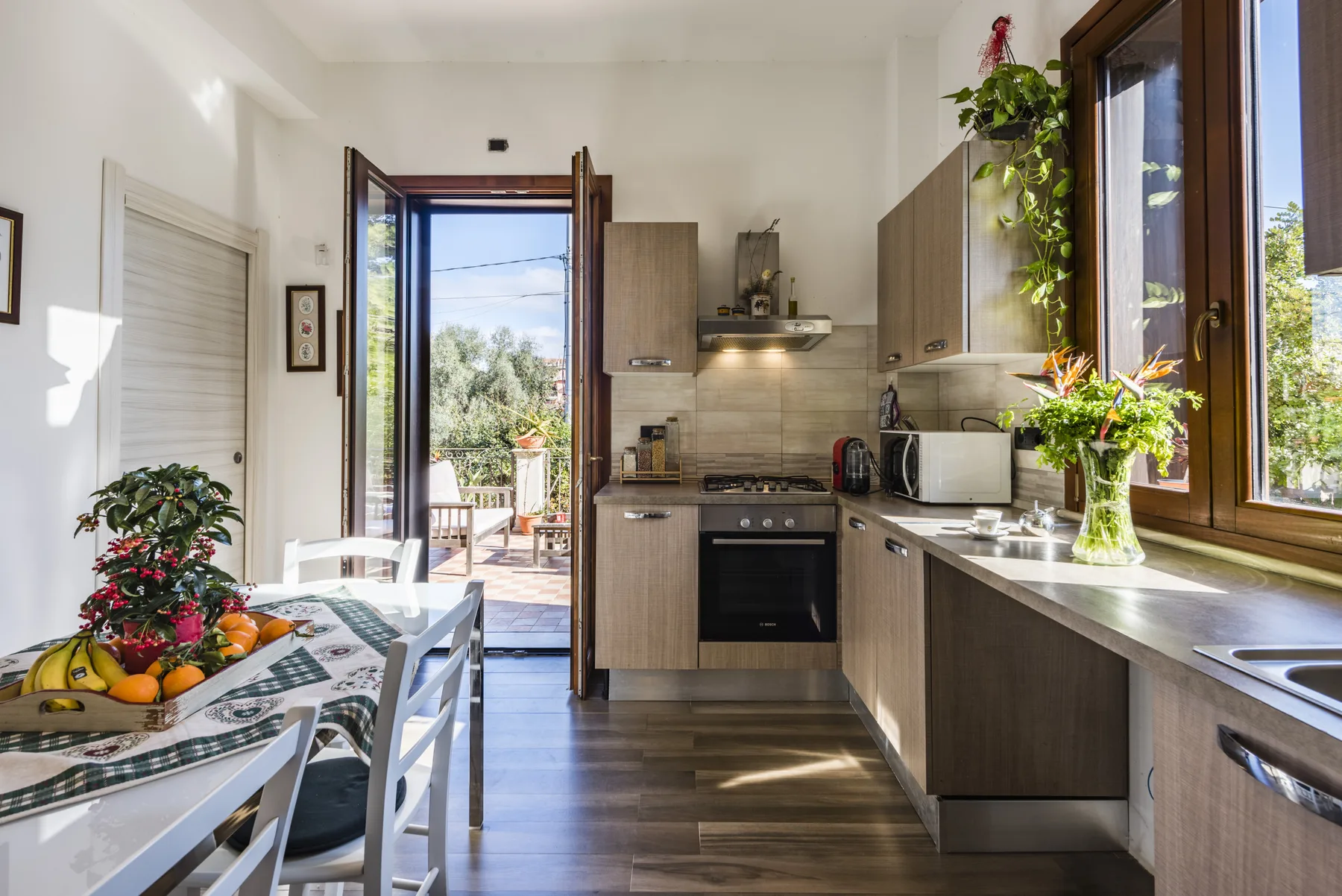 Una cocina y comedor luminosos con encimeras, armarios y una mesa junto a puertas abiertas.