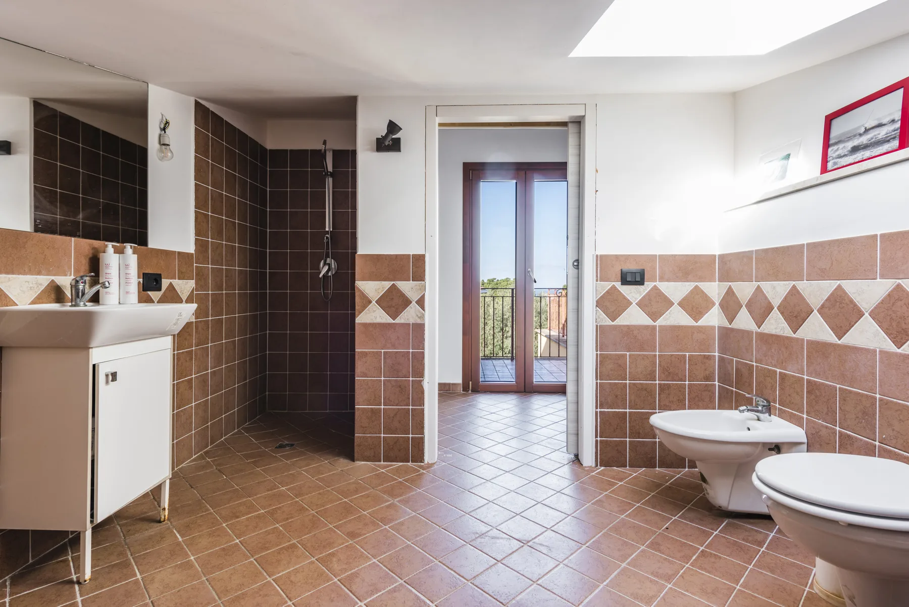 Un baño alicatado con lavabo, ducha e inodoros junto a una puerta de vidrio.