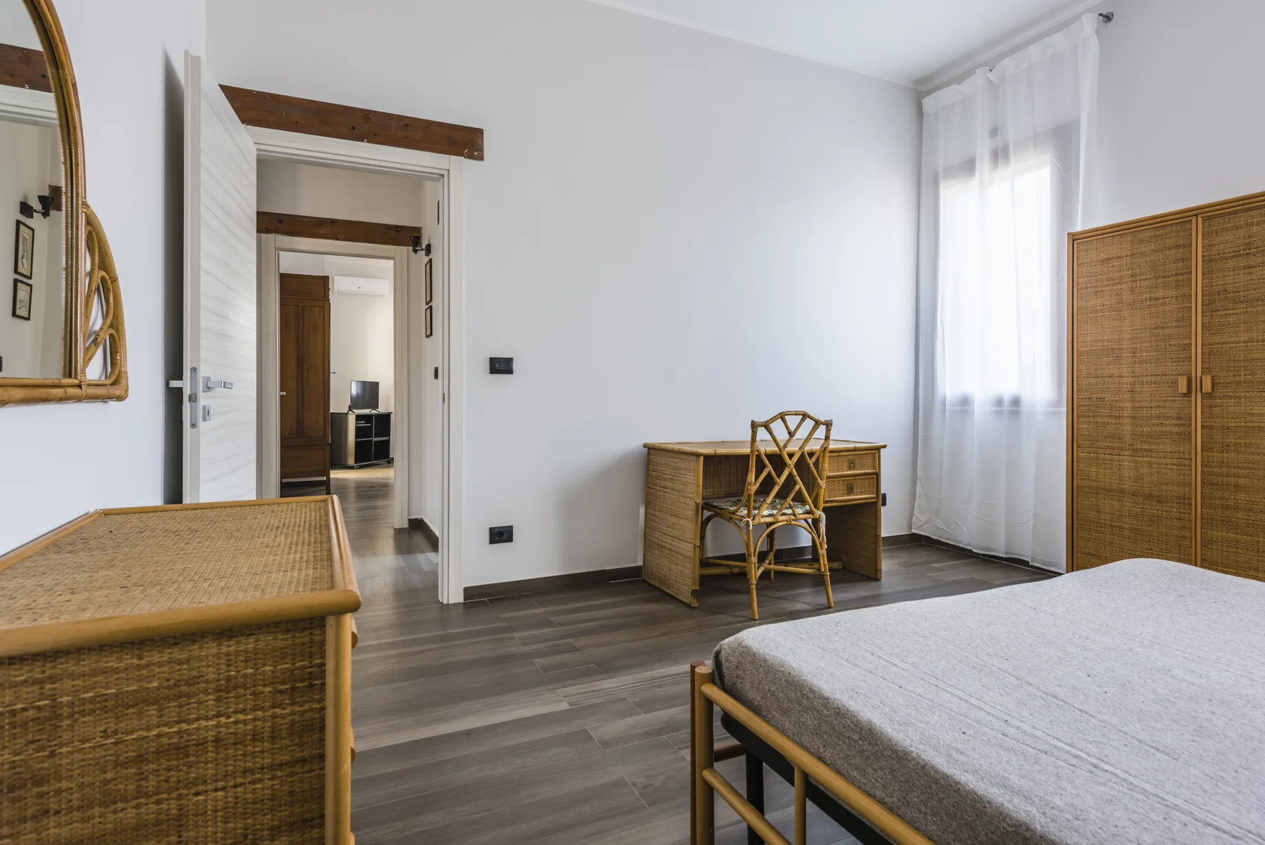 Un dormitorio luminoso con escritorio, asiento tejido y cama junto a un armario.