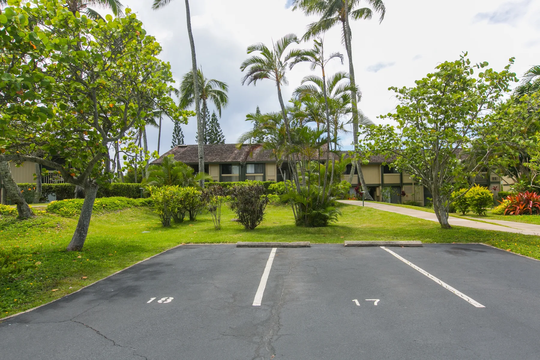 A landscaped residential building stands behind a paved parking area and palm trees.