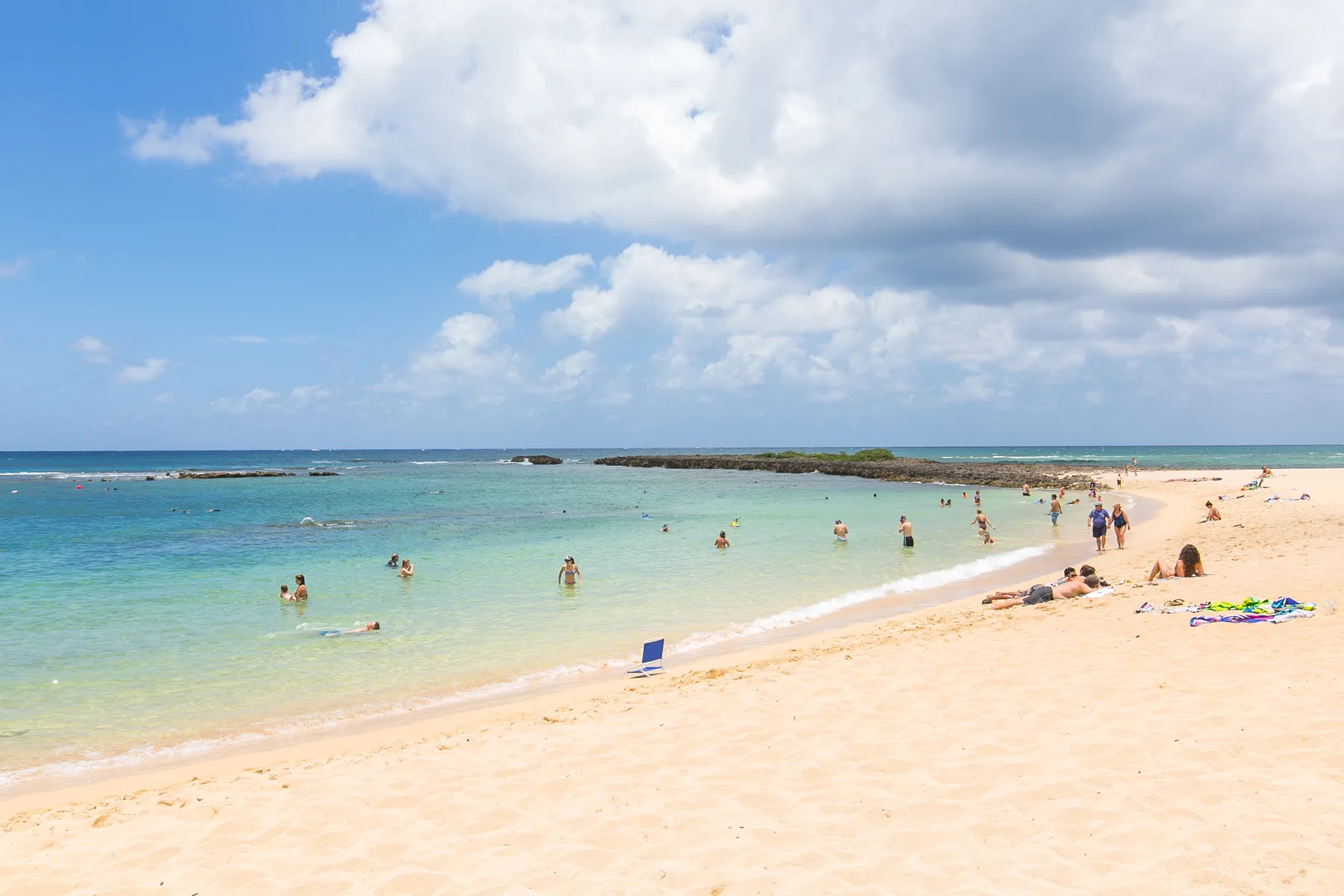 A sandy beach meets clear shallow water beneath a partly cloudy sky.