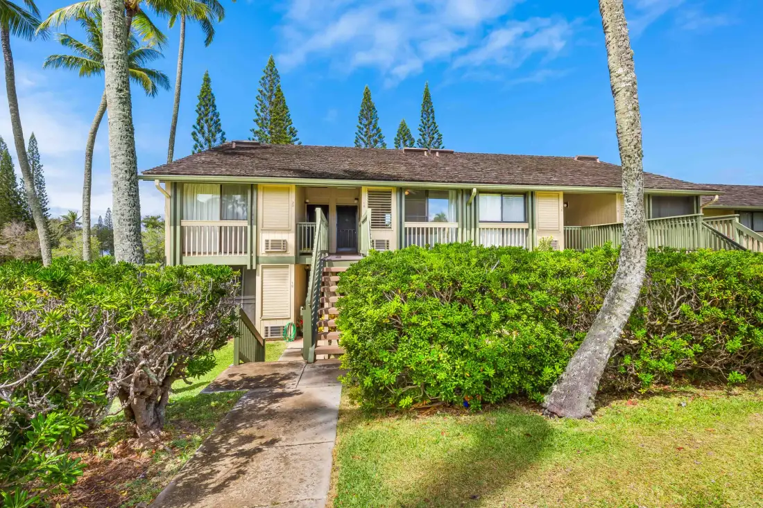 A low-rise building with a raised entrance stands behind green shrubs and palm trees.