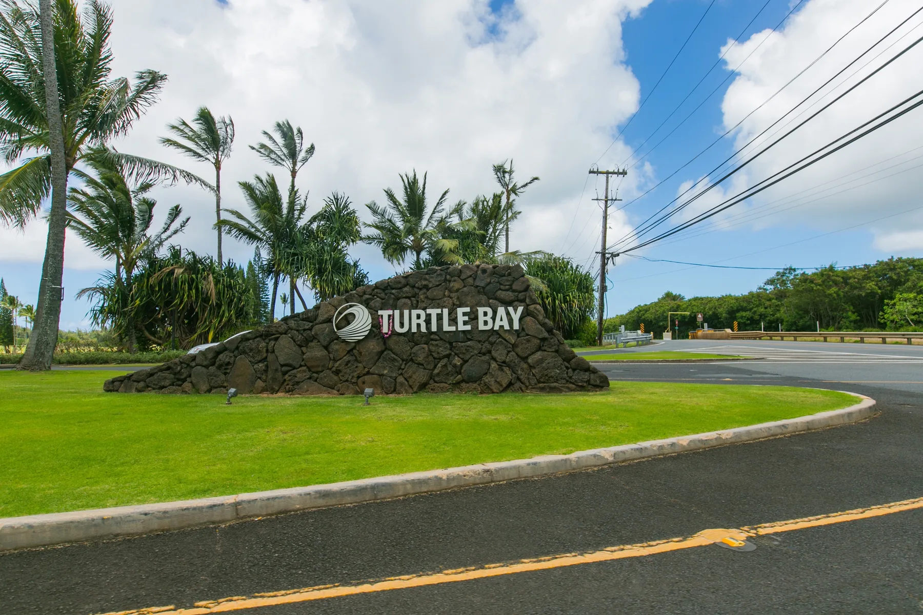 A roadside stone sign reading “Turtle Bay” sits beside a green lawn and palm trees.