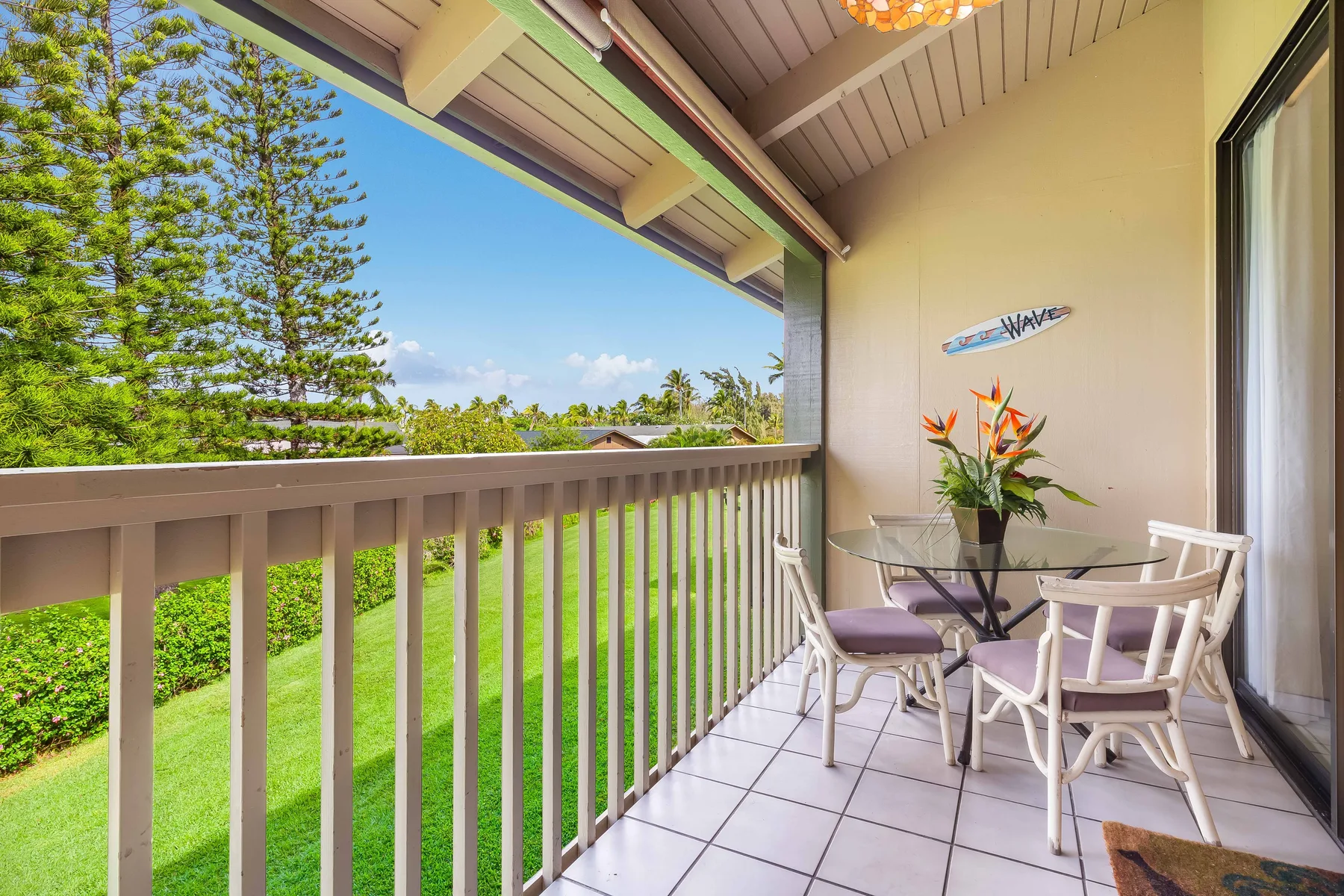 A covered balcony has a small table and seating overlooking green trees.