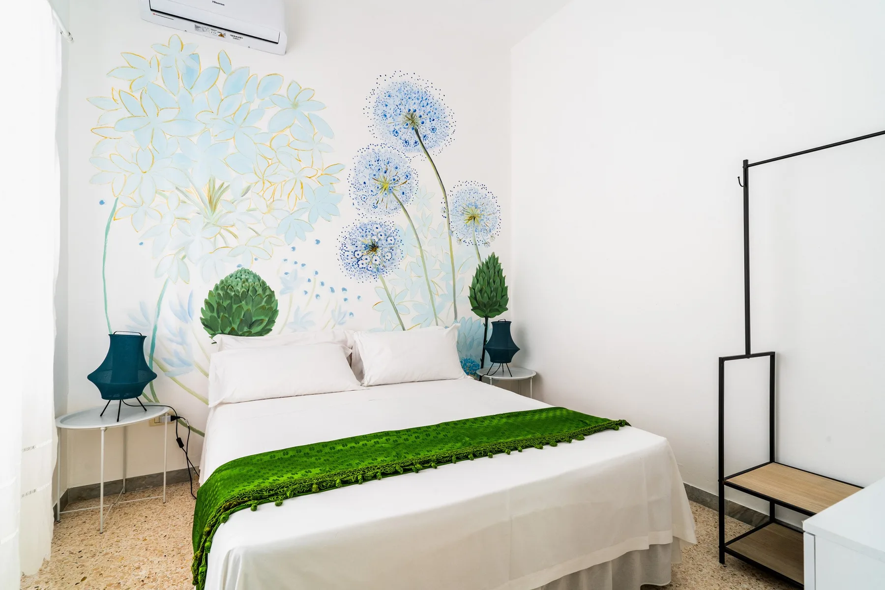 A bright bedroom features a white bed with a green cover and floral wall art.