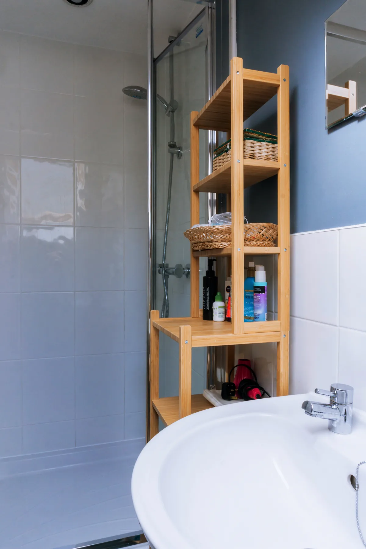 Un baño alicatado con ducha de vidrio, estantería de madera y lavabo blanco.