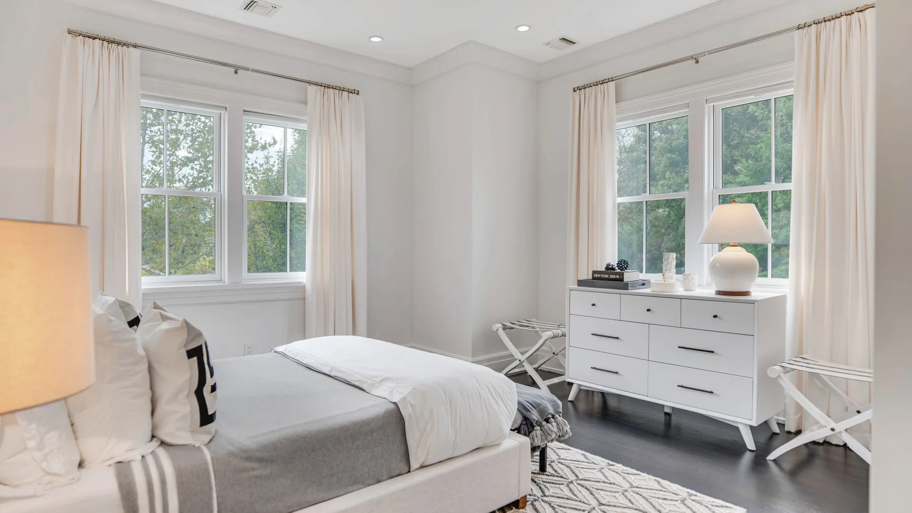 Un dormitorio luminoso con una cama arreglada y una cómoda blanca bajo las ventanas.
