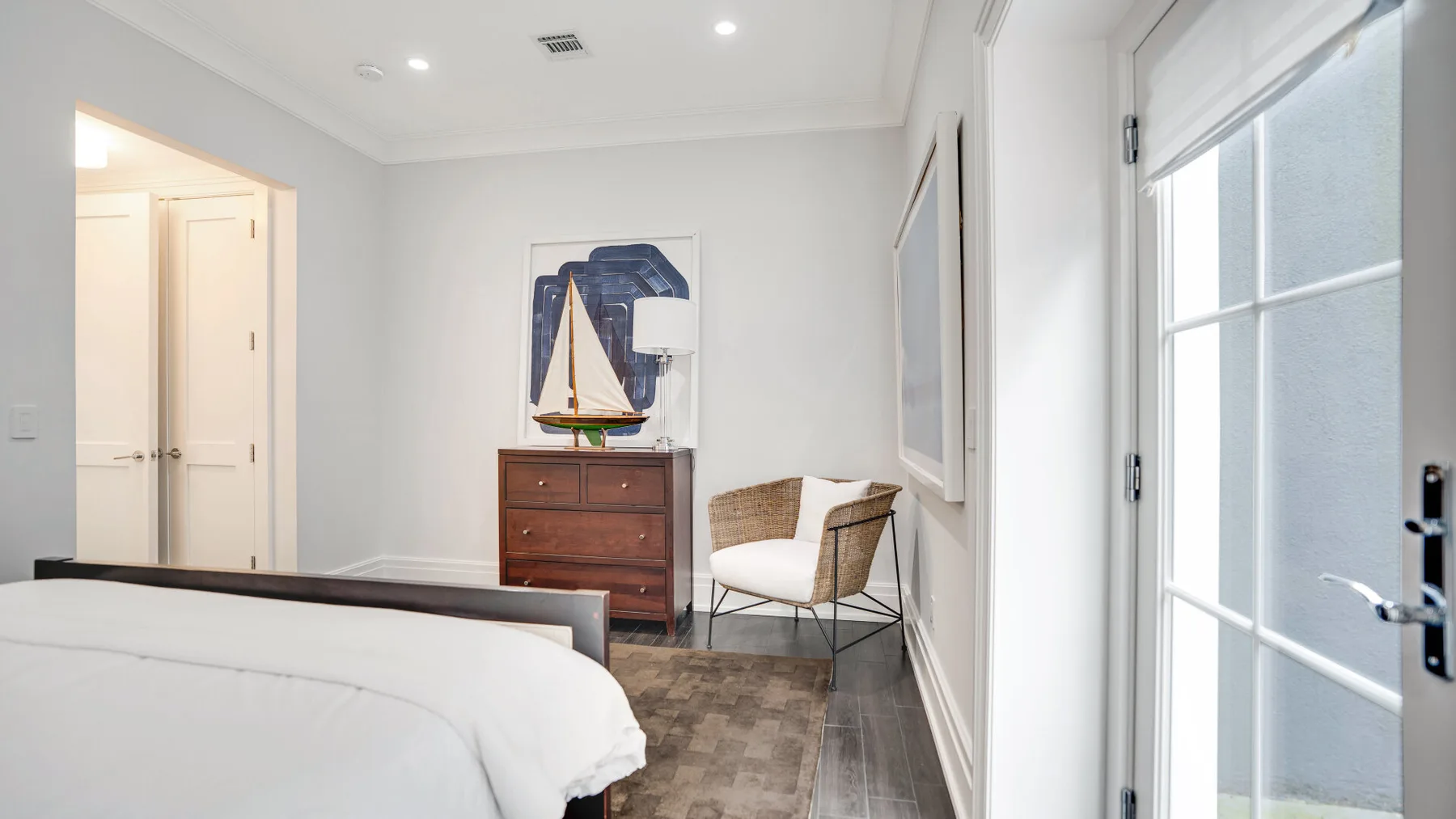 Un dormitorio luminoso con cama, cómoda de madera y asientos junto a la ventana.