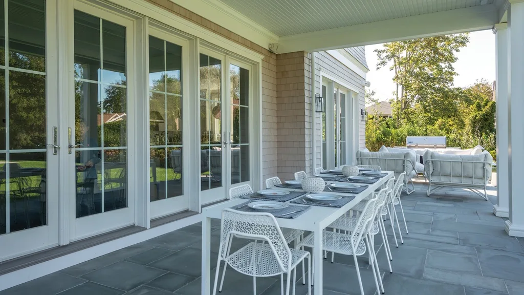 Un comedor exterior cubierto tiene una mesa larga y asientos blancos junto a puertas de vidrio.