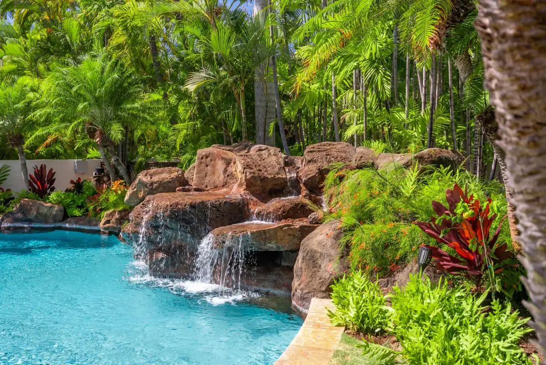 Una piscina tropical con agua cayendo sobre una formación rocosa, rodeada de vegetación exuberante.
