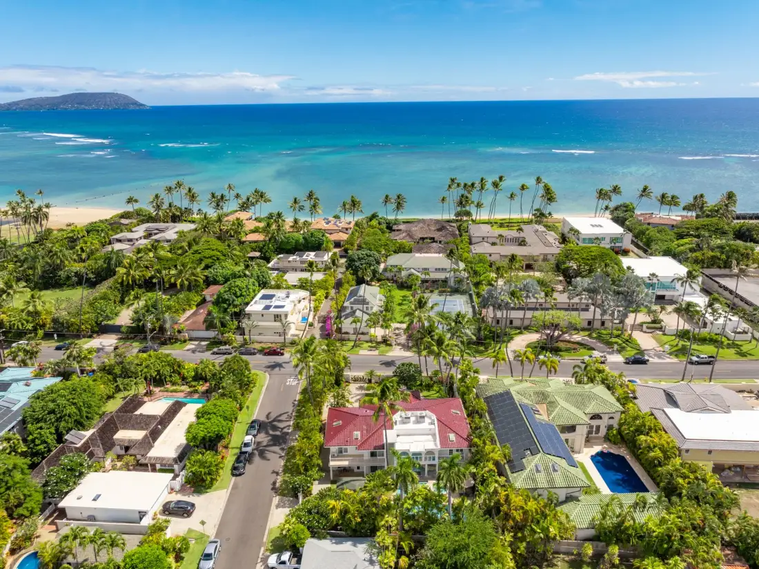 Vista aérea de un barrio costero tropical con casas, palmeras y el océano.
