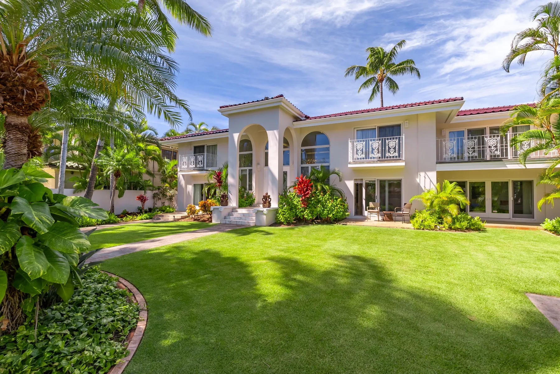 Una casa grande se alza detrás de un césped verde, rodeada de palmeras y vegetación tropical.