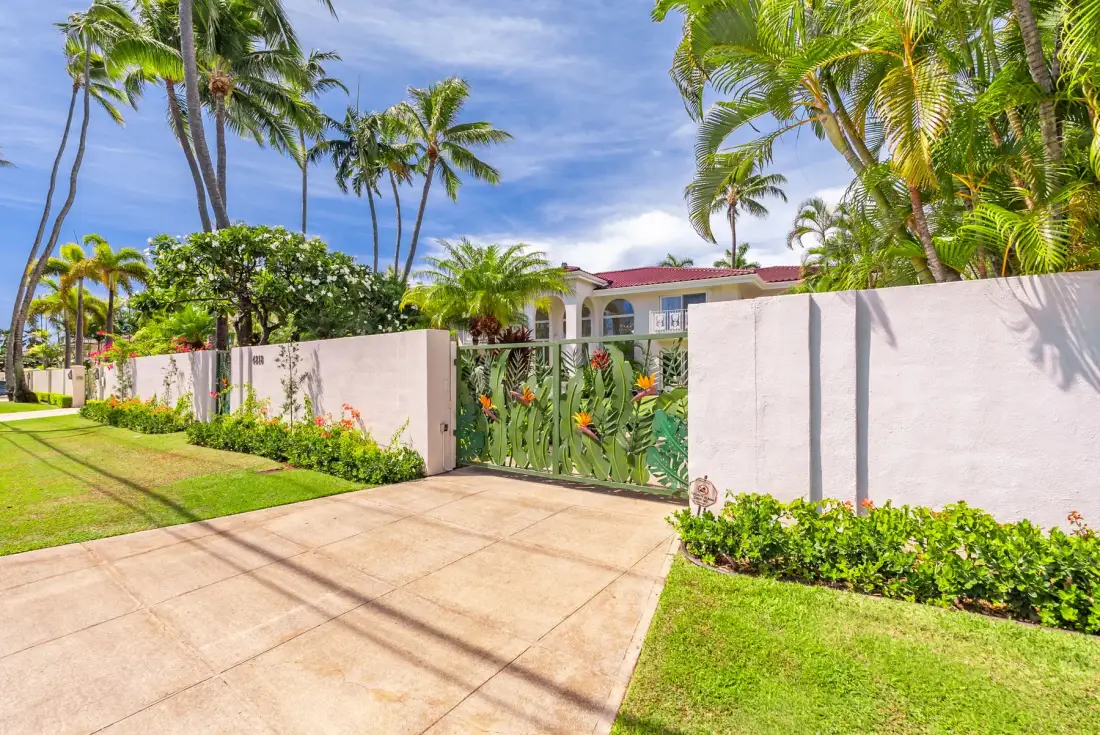 Una entrada con portón, rodeada por un muro blanco, plantas tropicales y palmeras.
